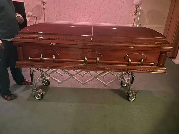 A polished wooden coffin rests on a wheeled metal cart in a room with soft pink wallpaper, creating a somber and respectful atmosphere.