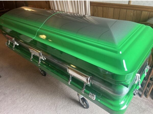 Green and silver casket on a wheeled cart in a room with wood-paneled walls and beige carpet. The casket has shiny silver handles.