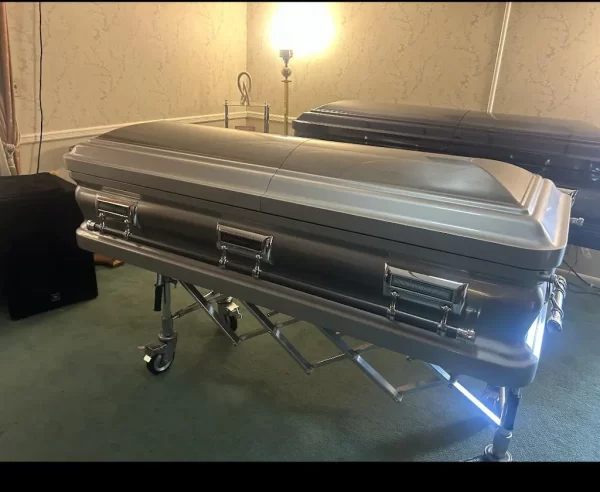 A silver metal casket on a trolley in a dimly lit room with light beige wallpaper and wooden trim. Another casket sits in the background.