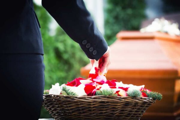 A person in a dark suit places flower petals into a basket beside a wooden coffin, conveying a solemn and respectful funeral atmosphere.