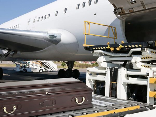 A polished brown coffin is being loaded onto a plane's cargo area at an airport. The scene conveys a solemn and respectful atmosphere.