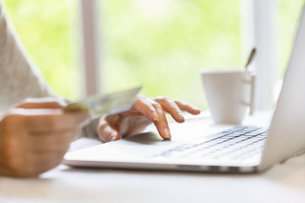 A person is using a laptop with one hand and holding a credit card in the other. A white coffee cup sits beside the laptop. The scene conveys online shopping.