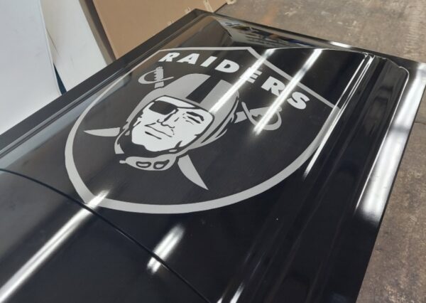 Black car hood displaying a Raiders logo, featuring a pirate with an eye patch inside a shield. The surface is glossy, reflecting overhead lights.