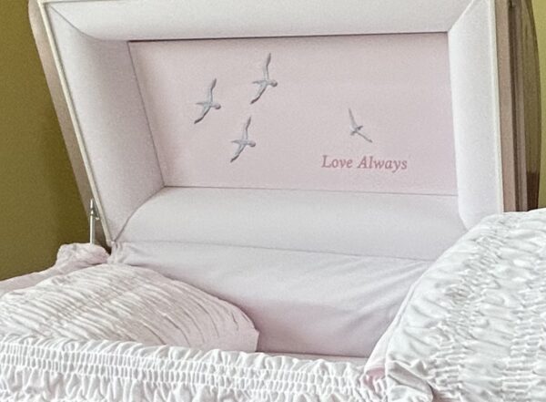 Open white casket with a cushioned interior, featuring flying birds and the words "Love Always" embroidered inside. The mood is serene and reflective.