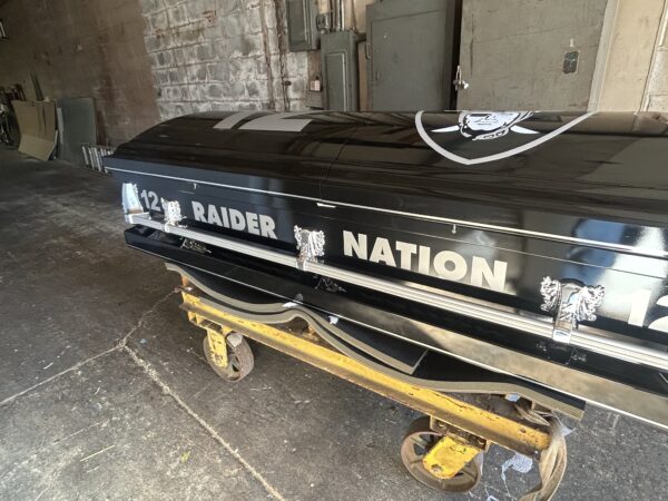 A black casket with "Raider Nation" and the number "12" in bold white lettering sits on a yellow metal trolley in a dimly lit room.