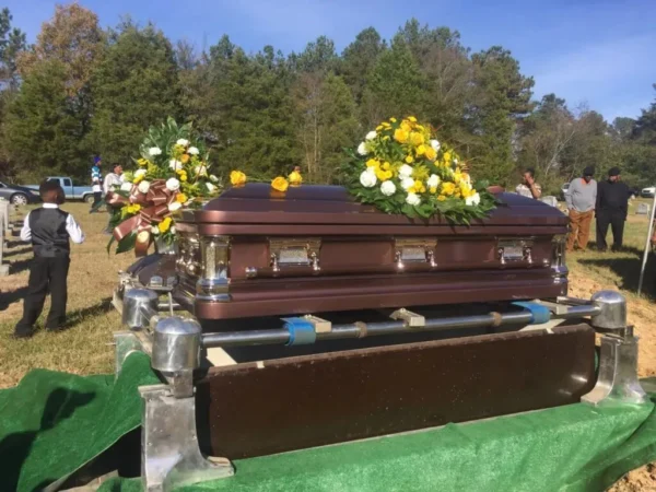 A brown casket adorned with yellow and white floral arrangements at a gravesite. People stand somberly nearby under a clear blue sky.