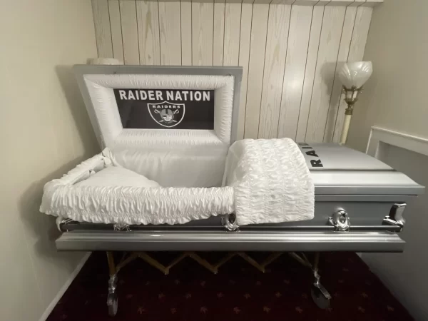 A silver casket adorned with "Raider Nation" branding inside the lid. The casket rests in a room with beige walls and a floor lamp, conveying a somber, yet personalized tribute.