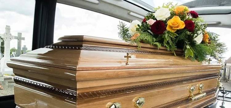 A polished wooden coffin adorned with red, yellow, and white flowers sits inside a hearse. The atmosphere appears solemn and respectful.