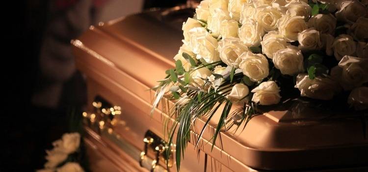 A closed bronze casket adorned with a large bouquet of white roses and greenery, conveying a solemn and respectful tone in a dimly lit setting.