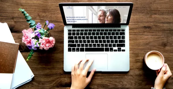 A laptop on a wooden table displaying a website, surrounded by a notebook, flowers, and a cup of coffee.