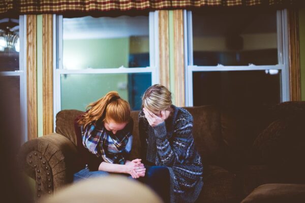 Two women sit on a couch in a dimly lit room, holding hands and bowing their heads. Their expressions appear somber and reflective, conveying a sense of support.
