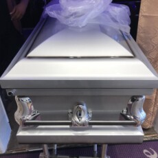 A silver casket with a smooth, shiny finish is adorned with chrome handles. A sheer white cloth rests on top. The scene conveys a somber and respectful atmosphere.