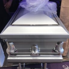 A silver casket with a smooth, shiny finish is adorned with chrome handles. A sheer white cloth rests on top. The scene conveys a somber and respectful atmosphere.