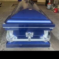 A glossy blue coffin with silver handles and ornate detailing sits on a concrete floor, surrounded by cleaning supplies in a garage-like setting.