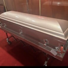 A closed, metallic pink coffin rests on a stand in a room with red carpet and brown-paneled walls, evoking a somber and respectful tone.