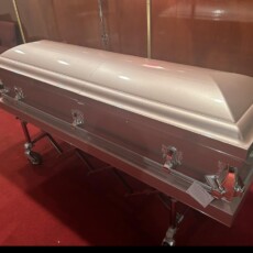 A closed, metallic pink coffin rests on a stand in a room with red carpet and brown-paneled walls, evoking a somber and respectful tone.