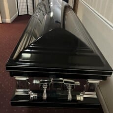 Sleek black coffin with reflective surface and silver handles, placed on maroon carpet in a dimly lit room, conveying a somber tone.