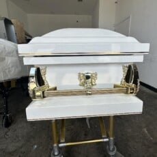 White casket with gold handles on a wheeled trolley in a plain room. The polished surfaces reflect light, creating a somber, respectful atmosphere.