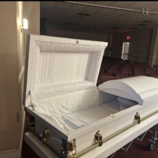 Open white casket with a white interior on a stand, located in an empty, dimly lit room with rows of stacked red chairs. The mood is solemn and quiet.