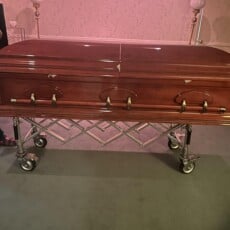 A polished wooden coffin on a wheeled stand in a softly lit room, conveying a somber and respectful tone. Handles and decorative details are visible.