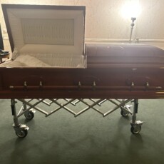 An open wooden casket on a wheeled stand in a softly lit room, creating a somber and respectful atmosphere. The casket's interior is lined with white fabric.