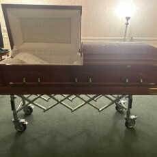 An open wooden casket on a wheeled stand in a softly lit room, creating a somber and respectful atmosphere. The casket's interior is lined with white fabric.