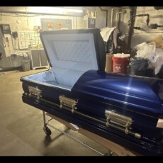 A shiny blue coffin with a light blue interior is open in a cluttered storage room. The setting is industrial, with pipes and warning signs visible.