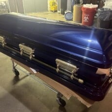 A glossy blue casket with silver handles on a wheeled cart in a dimly lit, cluttered room. The setting suggests a somber and industrial tone.
