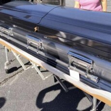 A shiny black metal casket rests on a wheeled stand outdoors. Two people partially visible nearby. Somber and reflective atmosphere.