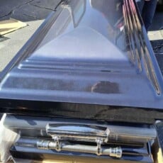 Black casket with shiny finish lying on pavement outside a vehicle. A person in a pink shirt stands nearby, creating a somber atmosphere.