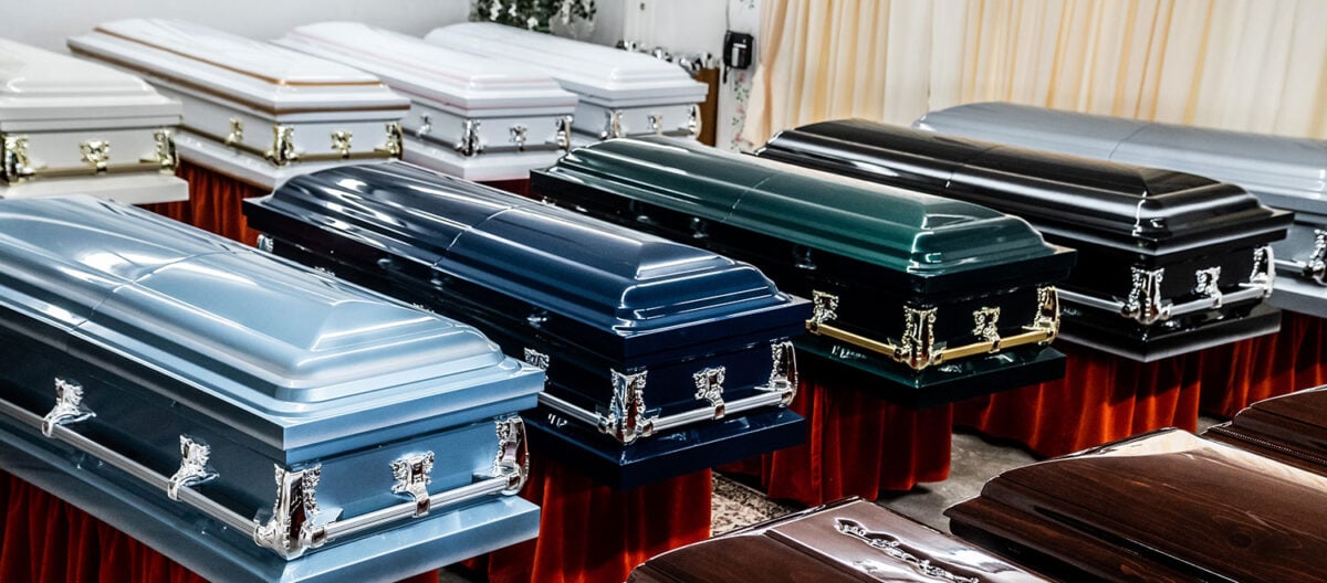 A display of ten closed caskets of varying colors, including blue, black, white, and brown, arranged neatly on red velvet stands in a well-lit room.