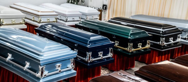 A display of ten closed caskets of varying colors, including blue, black, white, and brown, arranged neatly on red velvet stands in a well-lit room.
