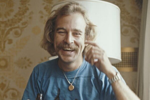 Smiling man with a mustache, wearing a blue shirt and necklace, sits casually in front of a lamp. The background shows patterned wallpaper. Warm and cheerful atmosphere.