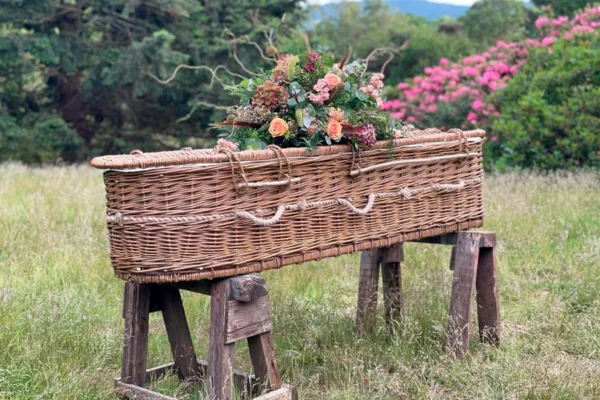 A wicker casket adorned with flowers rests on wooden supports in a grassy field. Pink flowering bushes and trees create a serene, natural backdrop.