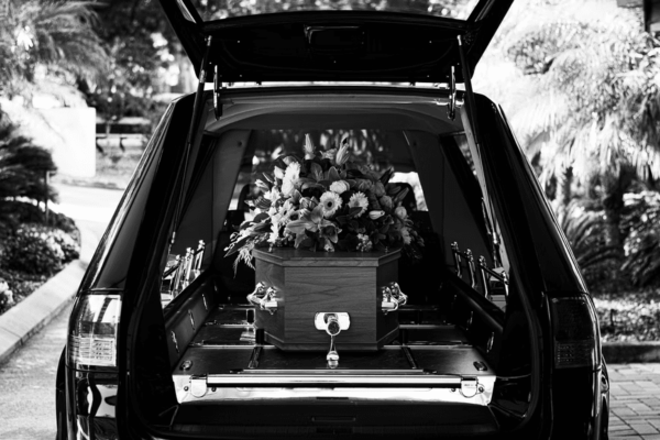 Black and white image of a hearse with an open back door. A casket adorned with a vibrant floral arrangement is inside, conveying a somber, respectful tone.