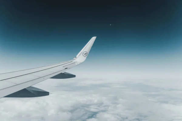 Airplane wing gliding above fluffy clouds with a clear blue sky in the background. A crescent moon is visible, adding a serene, peaceful tone.