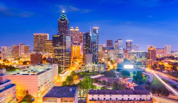 Vibrant cityscape of Atlanta at dusk, featuring illuminated skyscrapers against a vivid blue sky. Streets bustle with car lights, conveying urban energy.