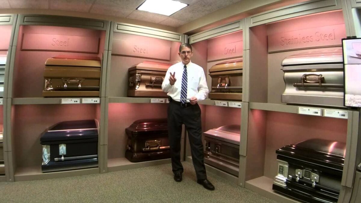 A person in formal attire stands in a showroom displaying various caskets. The lighting is warm, and the atmosphere is somber yet professional.