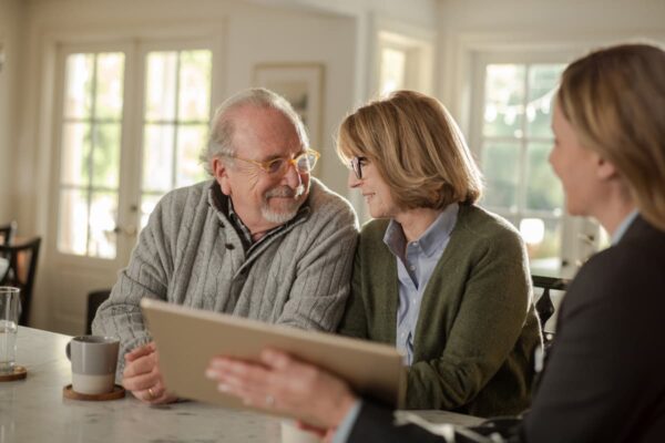 An elderly couple sits at a table, smiling at each other warmly. A professional with a clipboard engages them, creating a friendly, consultative atmosphere.