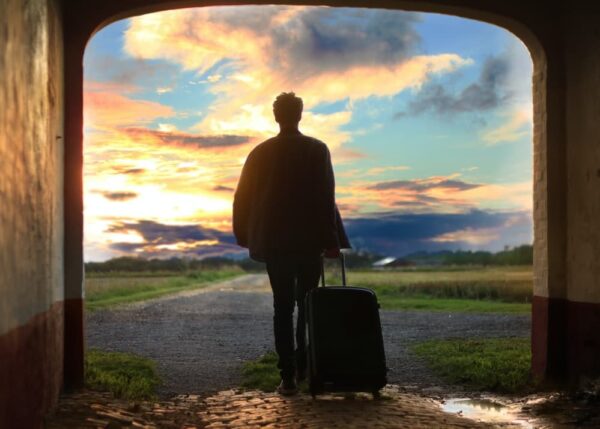 A person stands under an archway with a suitcase, gazing at a vibrant sunset over an open field. The scene conveys a sense of adventure and contemplation.