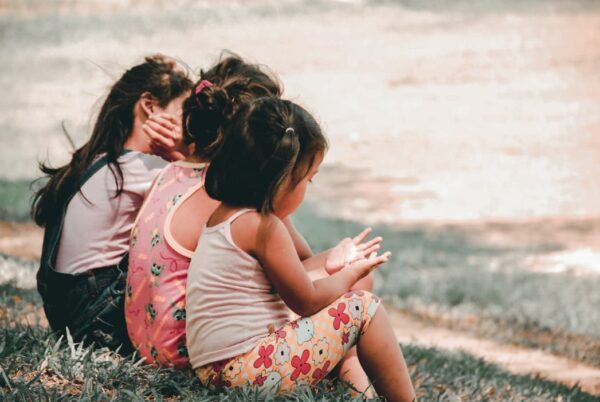 Three young girls sitting on grass, seen from behind, with one child holding something and another covering her face.