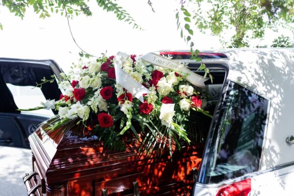 A wooden casket adorned with red and white floral arrangements is being placed into a hearse. The scene is shaded by trees, conveying a solemn tone.
