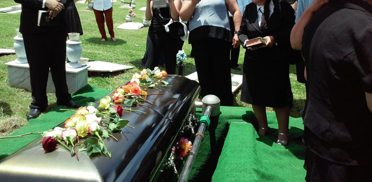 Closed casket with floral spray at a graveside service, surrounded by mourners dressed in black and standing on grass.