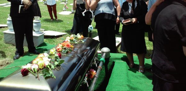 Closed casket with floral spray at a graveside service, surrounded by mourners dressed in black and standing on grass.