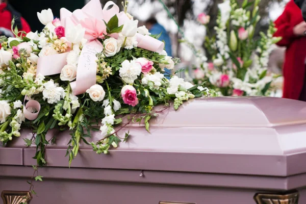 A lilac casket adorned with a bouquet of white and pink flowers, including roses and carnations, expressing a somber and respectful funeral setting.